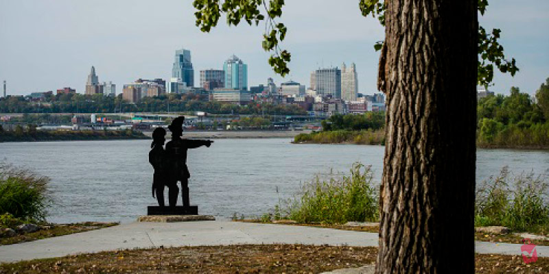 A silhouette statue of explorers points towards the Kansas City skyline across the water at Kaw Point Park.