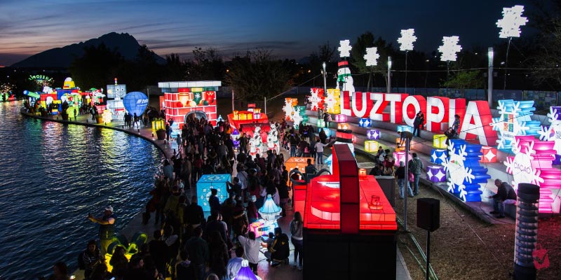 A vibrant night scene at Luztopía, a light festival, with numerous illuminated sculptures and displays along a body of water and people strolling through.