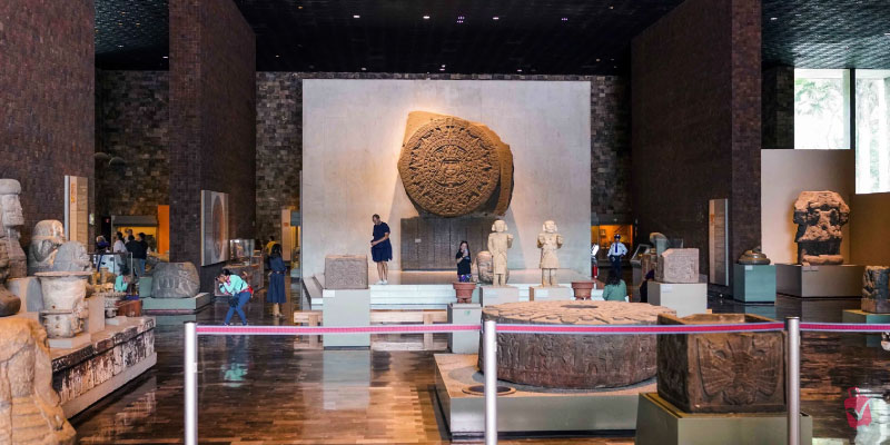 The interior of the Mexican History Museum, showcasing ancient stone artifacts and sculptures, including a large circular calendar stone.