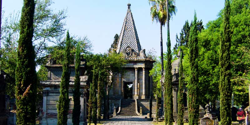 The ornate entrance to Museo Panteón de Belén, an ancient cemetery in Guadalajara, surrounded by tall trees and lush greenery.