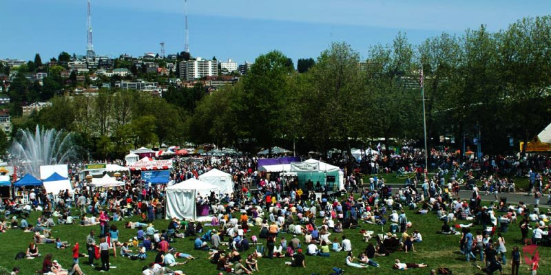 Crowds gather on a sunny day at the Northwest Folklife Festival in a park with tents and a fountain in the background.