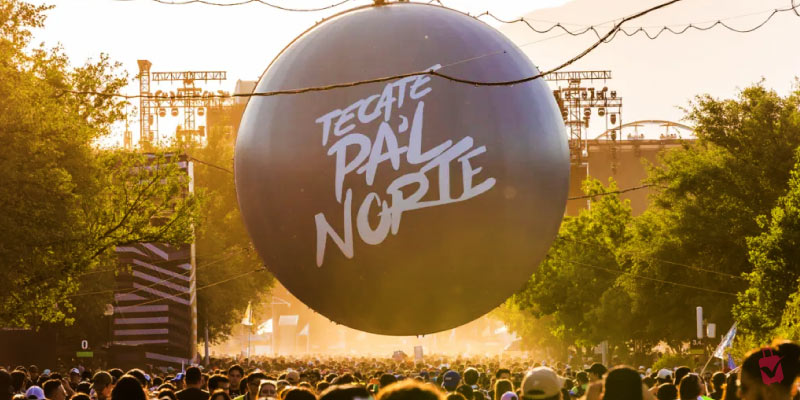 A large "TECATE PA'L NORTE" balloon suspended over a huge crowd at an outdoor music festival during the day.