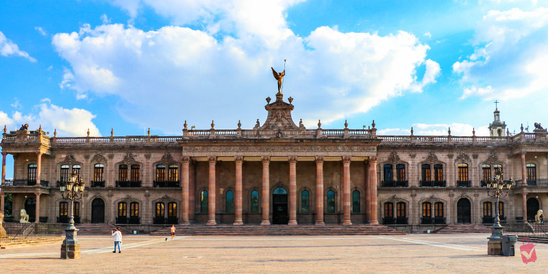 The Palace Museum in Monterrey, Mexico, a large, ornate building with many columns and a statue on the roof, under a blue sky with clouds.