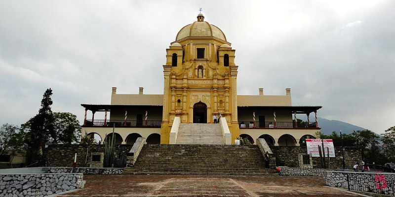 The Palacio del Obispado in Monterrey, Mexico, a grand yellow building with a large staircase and archways, under a cloudy sky.