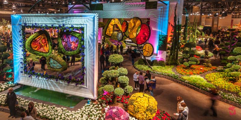 A vibrant display of intricate floral arrangements and garden designs fill a large exhibition hall at the Philadelphia Flower Show, with numerous visitors admiring the exhibits.