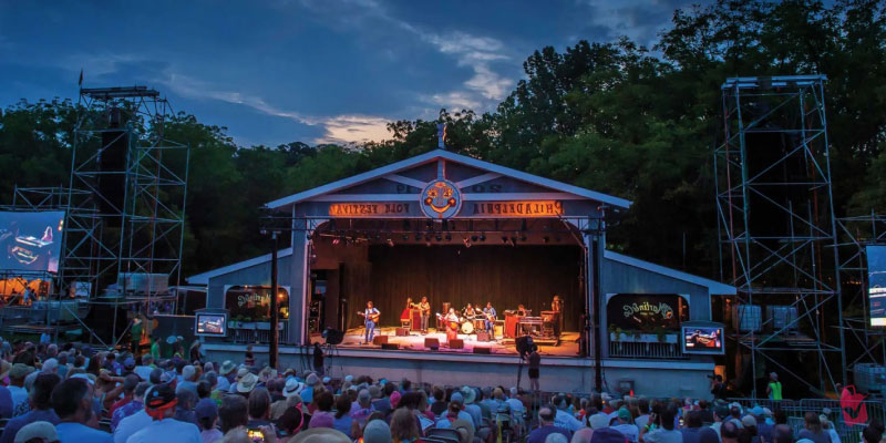 An audience gathers before an outdoor stage where a band performs at the Philadelphia Folk Festival.