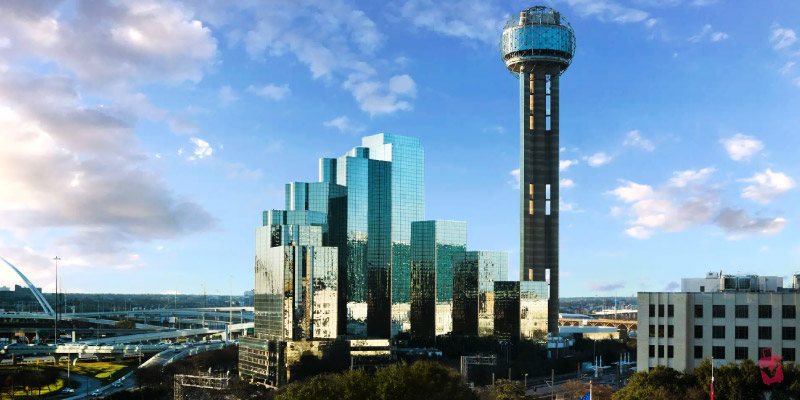 Modern skyscrapers stand next to the iconic Reunion Tower with its spherical observation deck under a bright blue sky with light clouds.