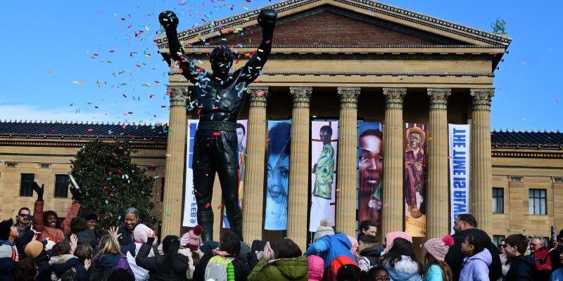 A large crowd gathered in front of the Philadelphia Museum of Art, celebrating RockyFest 50 with confetti falling around the Rocky statue.