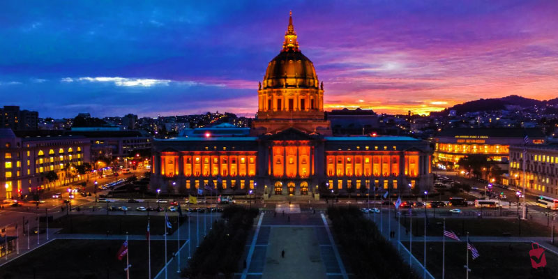 San Francisco City Hall glows with golden lights at dusk featuring its grand dome and illuminated facade against a vibrant sunset sky.