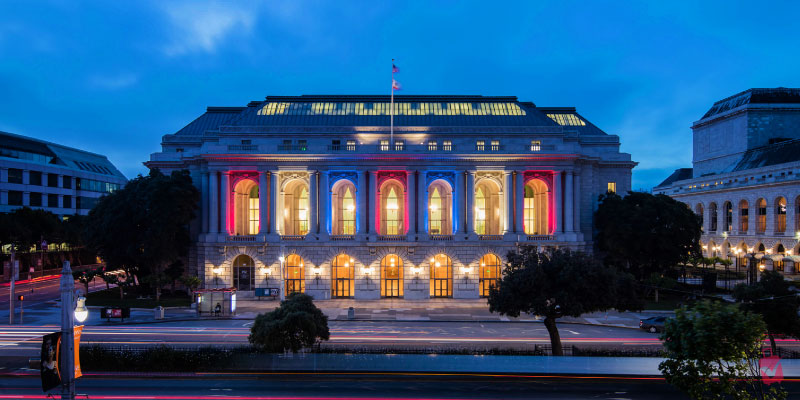 The San Francisco War Memorial and Performing Arts Center shines at night with red, white, and blue lights highlighting its classical architecture.