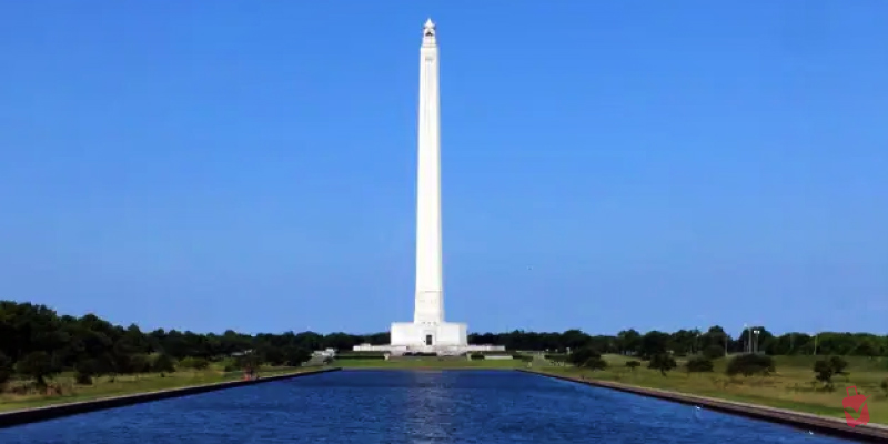 The towering San Jacinto Monument stands over a long reflecting pool under a clear blue sky and represents the San Jacinto Museum historical site.