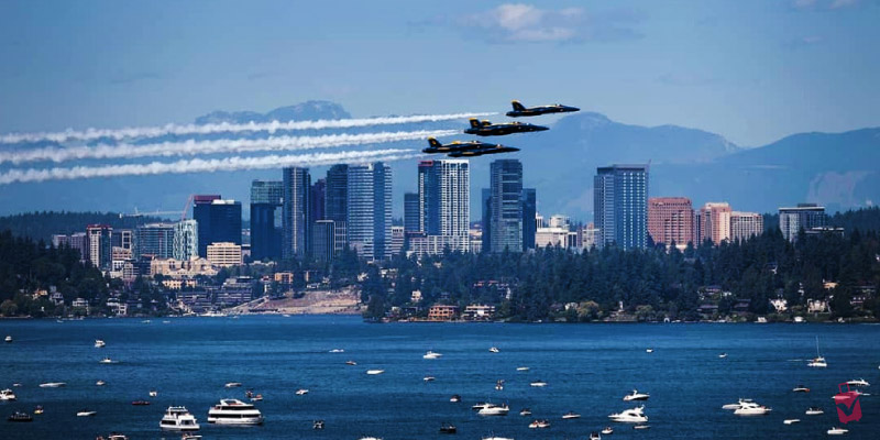 Three fighter jets from the Seafair Weekend Festival fly in formation over a city skyline and a body of water filled with boats.