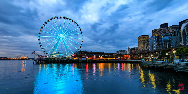 The Seattle Great Wheel glows with blue lights at dusk, reflecting in the water, with the city skyline behind it.