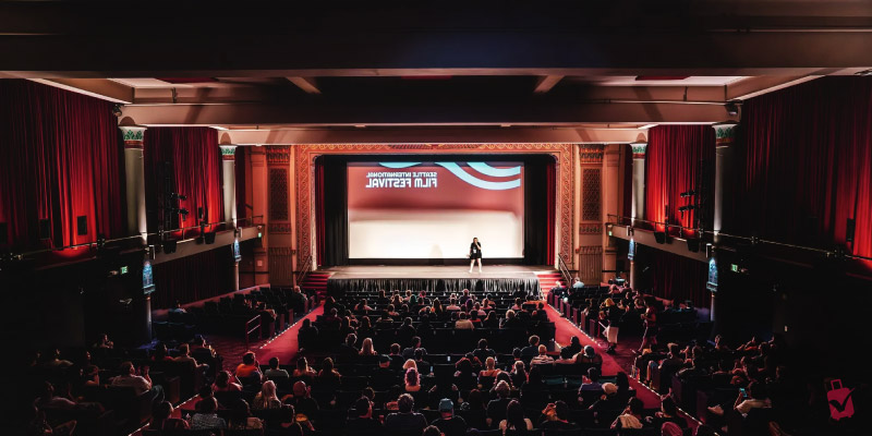 A large audience watches a speaker on stage at the Seattle International Film Festival in a historic theater with red curtains.