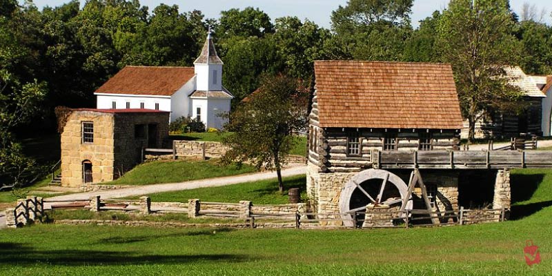 The Shoal Creek Living History Museum showcases a rustic village with a log cabin, waterwheel, and white church.