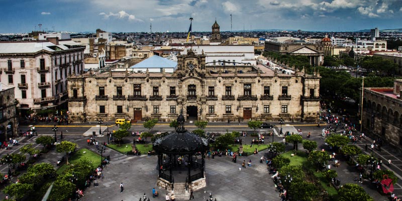 The State of Jalisco Government Building in Guadalajara, a stately stone structure, overlooking a bustling public square with a gazebo.