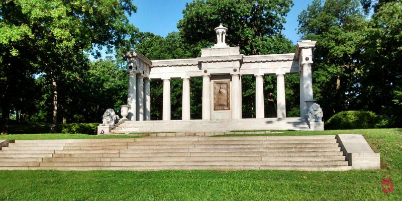 A grand stone structure with columns and steps is nestled among lush green trees in Swope Park.