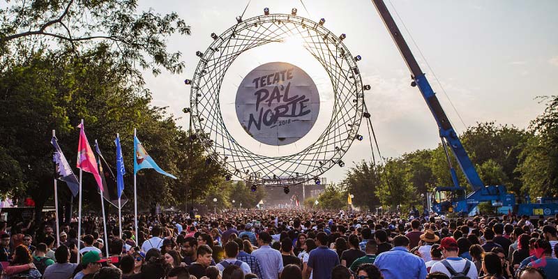 A massive crowd gathers under a large, circular stage structure with the words Tecate Pa'l Norte in the center, surrounded by trees.