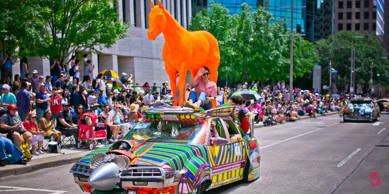 A colorful car topped with a giant orange horse drives through crowds of spectators during the creative and unique Houston Art Car Parade.