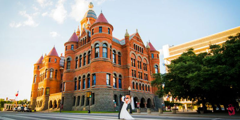 A wedding couple poses in front of the historic red sandstone and granite architecture of The Old Red Courthouse under a clear sky.