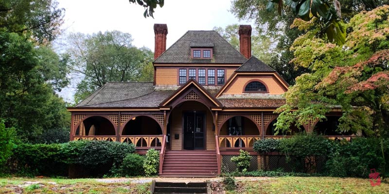 The Wren’s Nest is a historic Victorian-style cottage with a wrap-around porch and decorative gables surrounded by lush green trees.