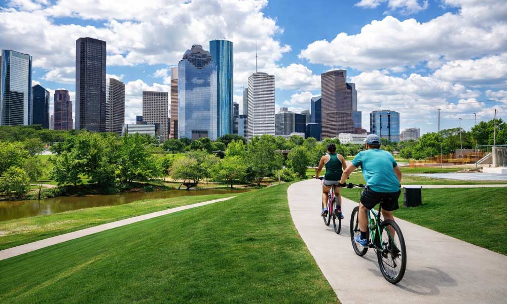 Two cyclists enjoy a scenic ride on a paved path in Buffalo Bayou Park with the downtown skyline in the distance as part of the many active things to do in houston.