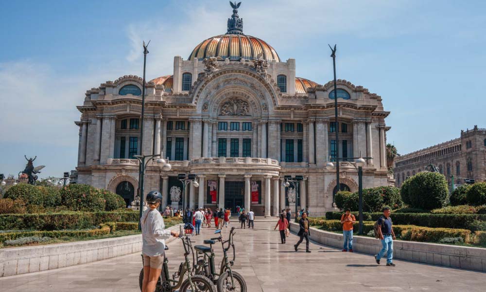 A sunny day at the Palacio de Bellas Artes, with people walking and biking around the grand architectural landmark, showcasing things to do in Mexico City.