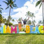 A couple jumping in front of a colorful "MIAMI BEACH" sign with palm trees and buildings in the background.