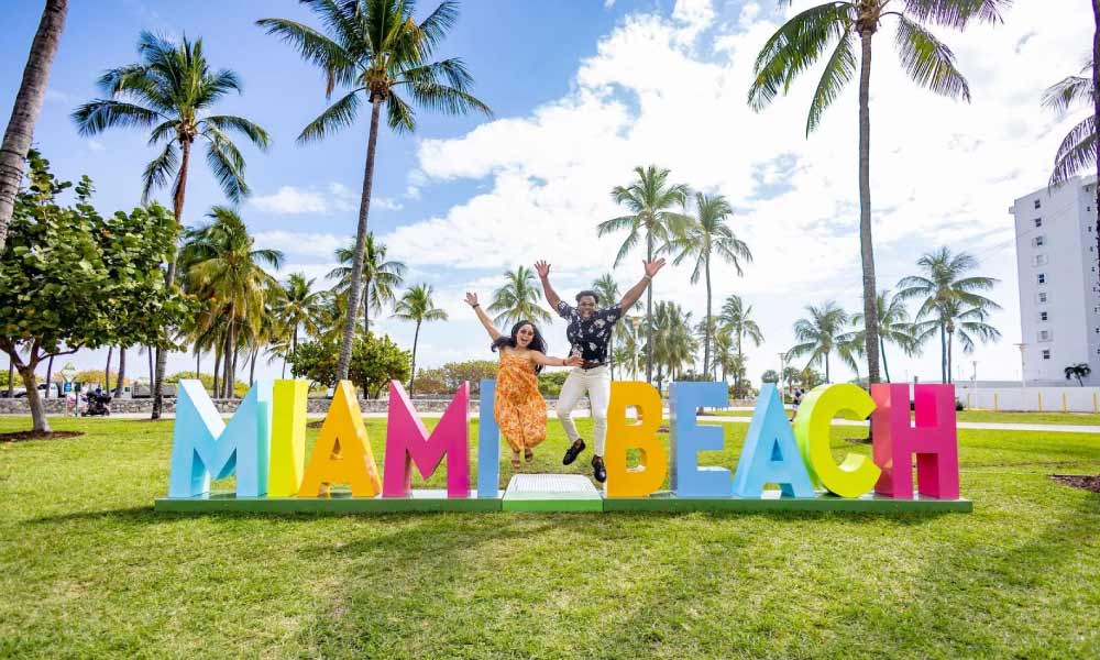A couple jumping in front of a colorful "MIAMI BEACH" sign with palm trees and buildings in the background.