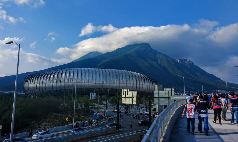 People walk on a bridge towards a large, modern stadium with a distinctive roof, set against a backdrop of majestic mountains under a blue sky with some clouds.