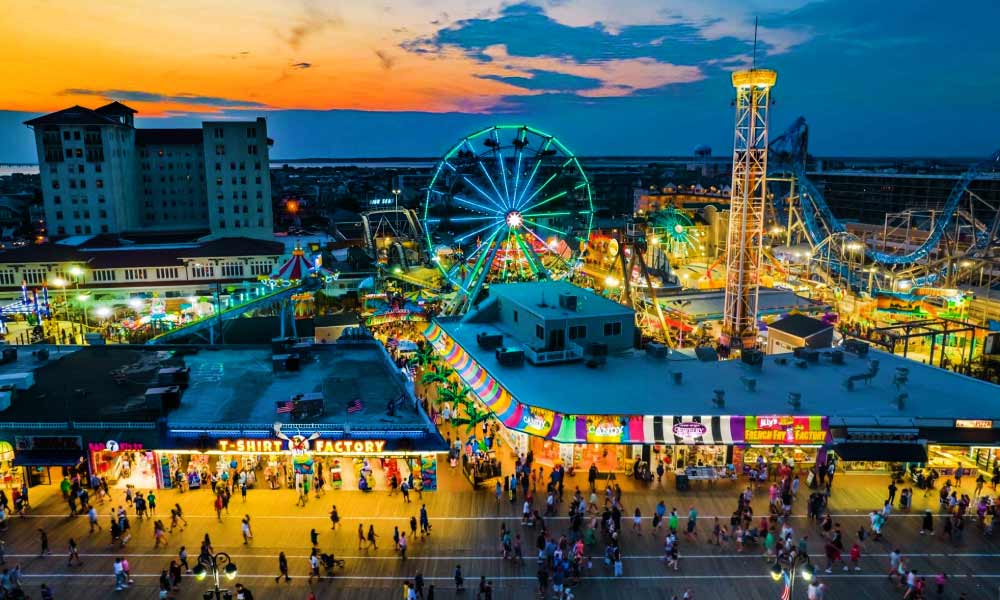 Bright nighttime view of an amusement park boardwalk with a glowing Ferris wheel and shops representing popular Things to Do in New Jersey.