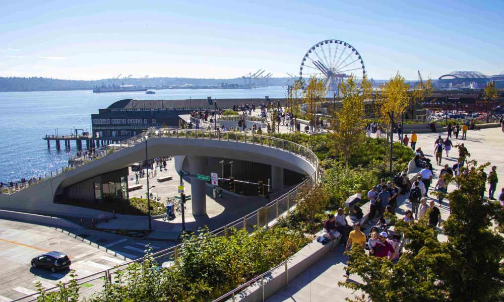 People walk along a pedestrian bridge and the Elliott Bay waterfront, highlighting popular things to do in Seattle like visiting the Great Wheel.