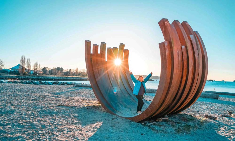 A girl poses inside a large metal arc sculpture at Sunset Beach with the sun shining through, showcasing popular things to do in Vancouver.