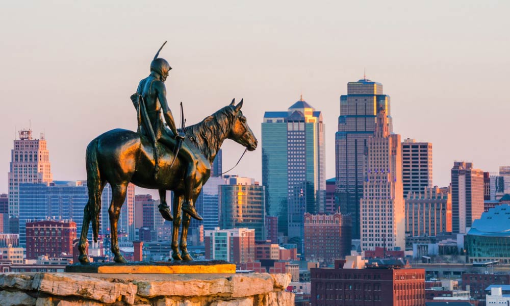 A statue of a Native American on horseback overlooks the Kansas City skyline at sunset.