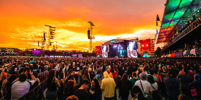 A large outdoor music festival at sunset with a huge crowd and a brightly lit stage under an orange sky.