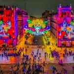 A city square at night illuminated with colorful projections of skeletons and floral designs on the surrounding buildings, with many people below featuring the festivals and events in Mexico City.