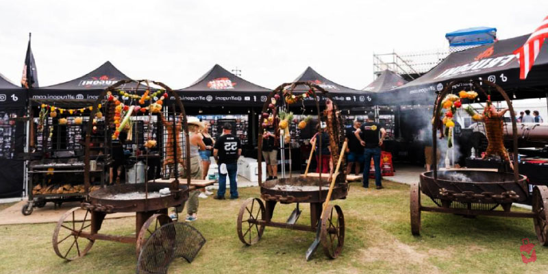 Iron rotisserie grills with hanging meats and pineapples at the World’s Championship Bar-B-Que Contest as visitors gather to sample smoked treats.