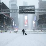 A person walks with a suitcase on a snow-covered Times Square street, empty due to a Travel Ban in NYC from a Winter Storm. Buildings with billboards stand in the falling snow.