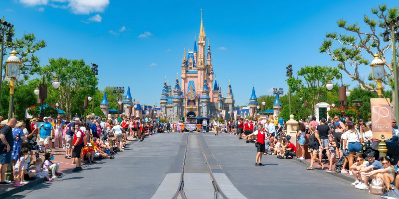 Large crowds of visitors lining Main Street, U.S.A., watching a parade with Cinderella Castle in the background under a clear blue sky.